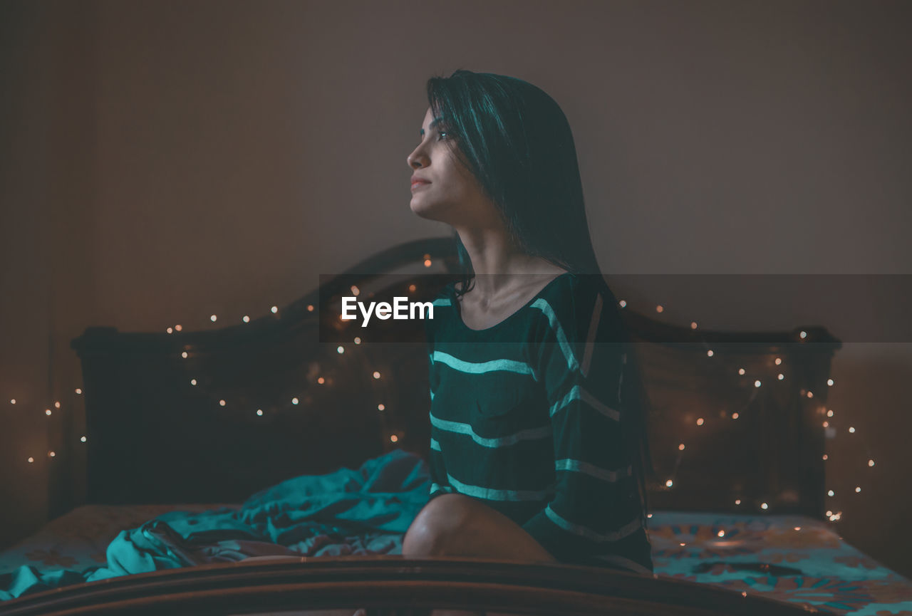 YOUNG WOMAN SITTING ON BED IN BEDROOM