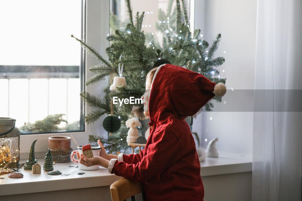 A thoughtful boy in a santa costume sits at the window and makes christmas decorations