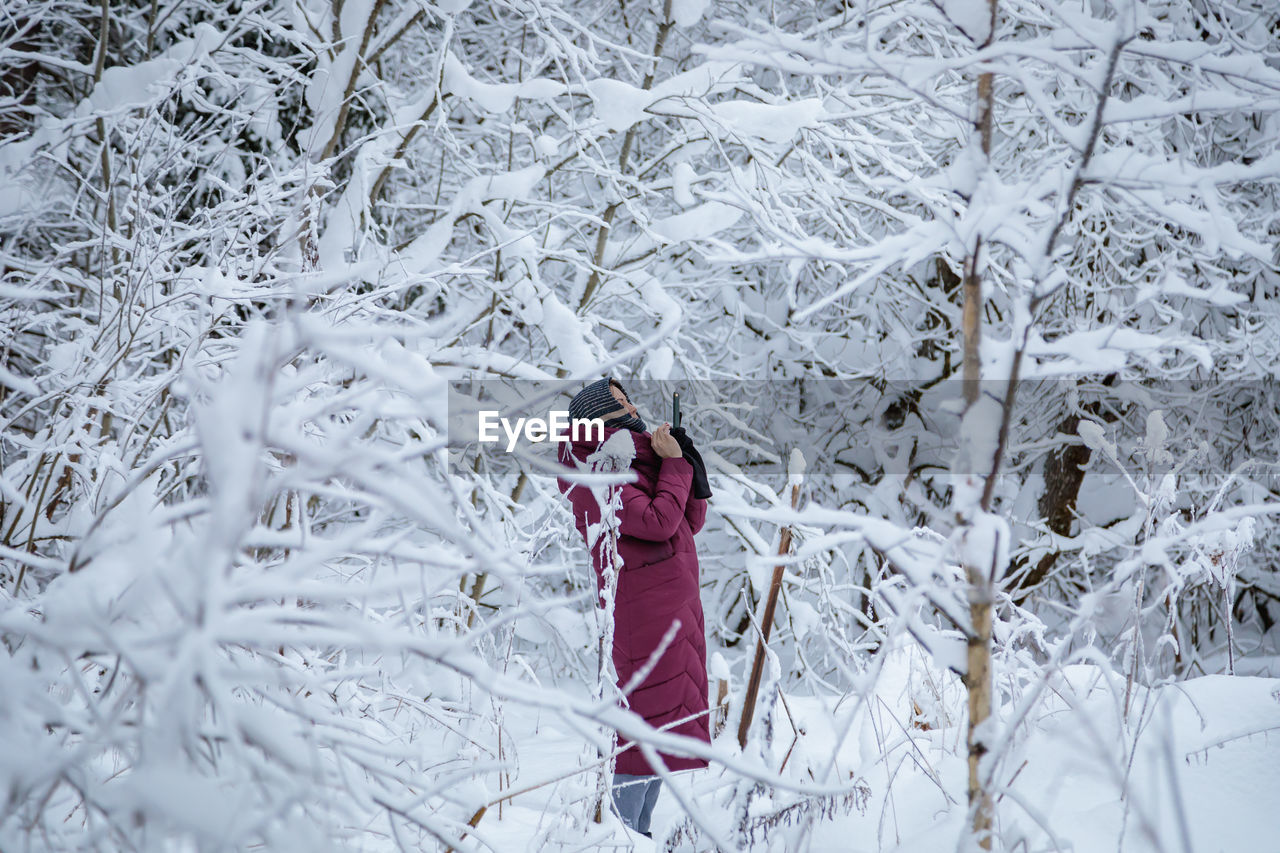 A girl in a burgundy down jacket photographs a snow-covered forest on a mobile phone. person