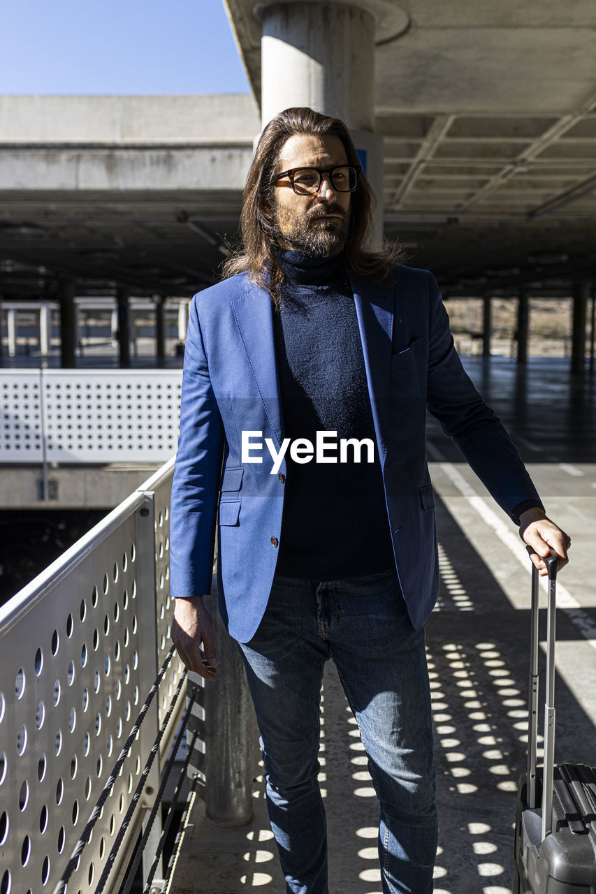 Handsome trendy unshaven adult male in stylish outfit and eyeglasses looking away while standing with suitcase on airport terrace in sunny day