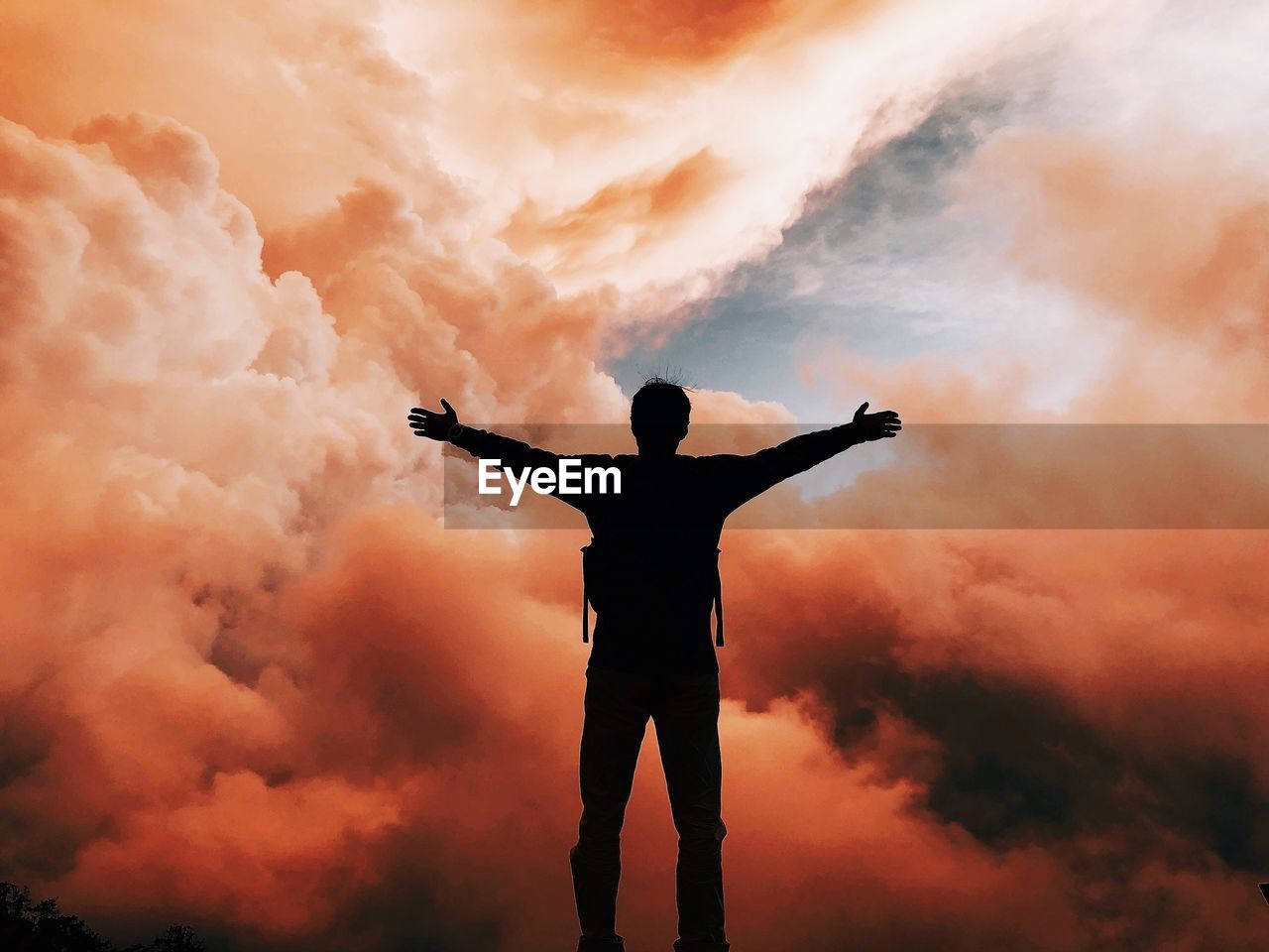 Man with arms outstretched standing against cloudy sky during sunset