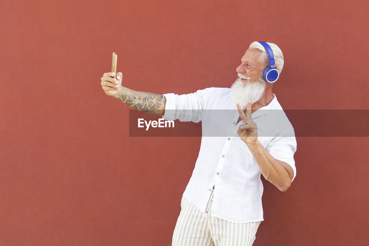 Smiling man taking selfie and listening to music while standing against wall