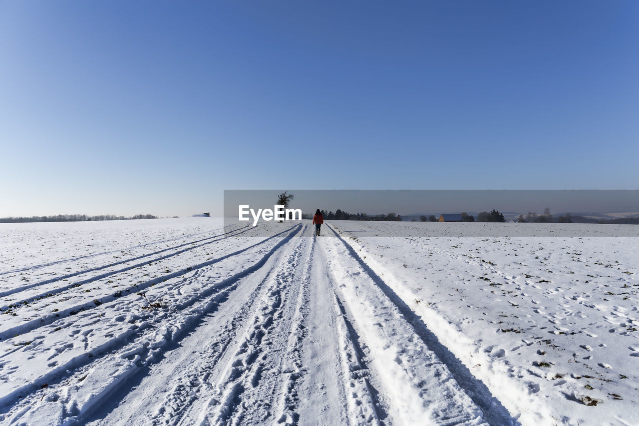 Rear view of person walking on snow covered field against clear blue sky