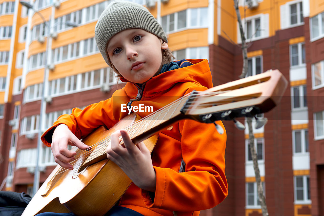 Portrait of caucasian blond boy in orange jacket playing acoustic guitar outdoors on autumn day