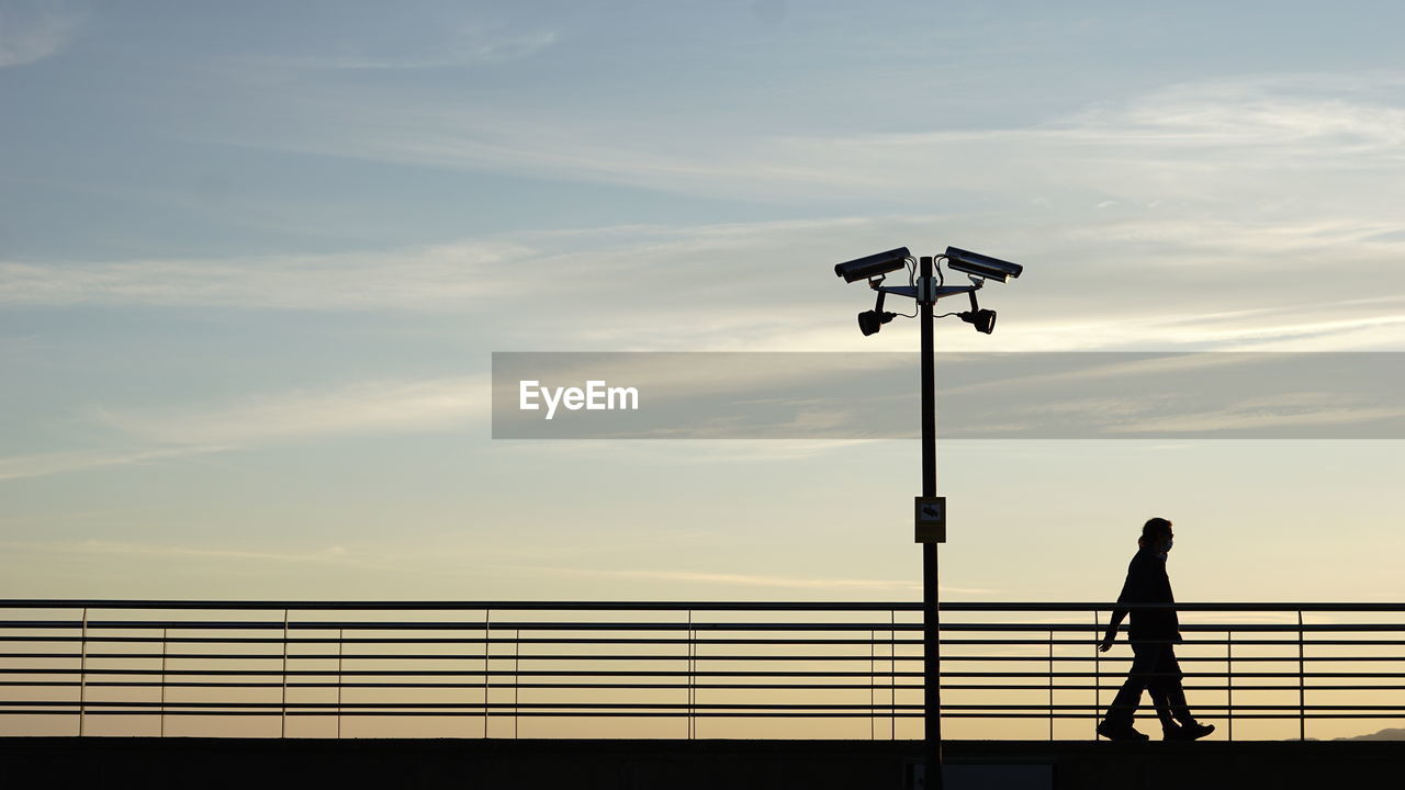 Silhouette man standing by railing and cctv against sky