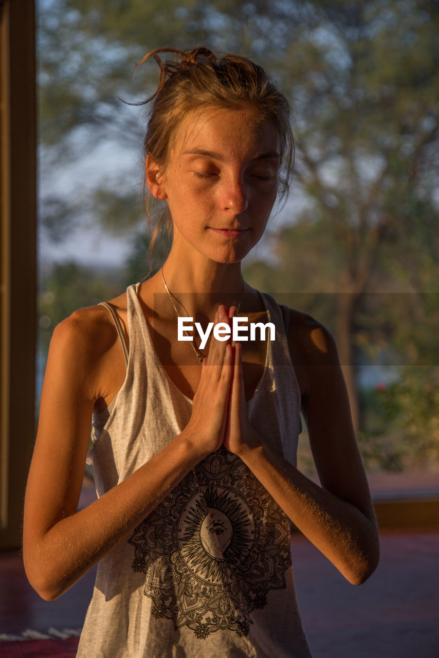 Teenage girl meditating against window during sunset