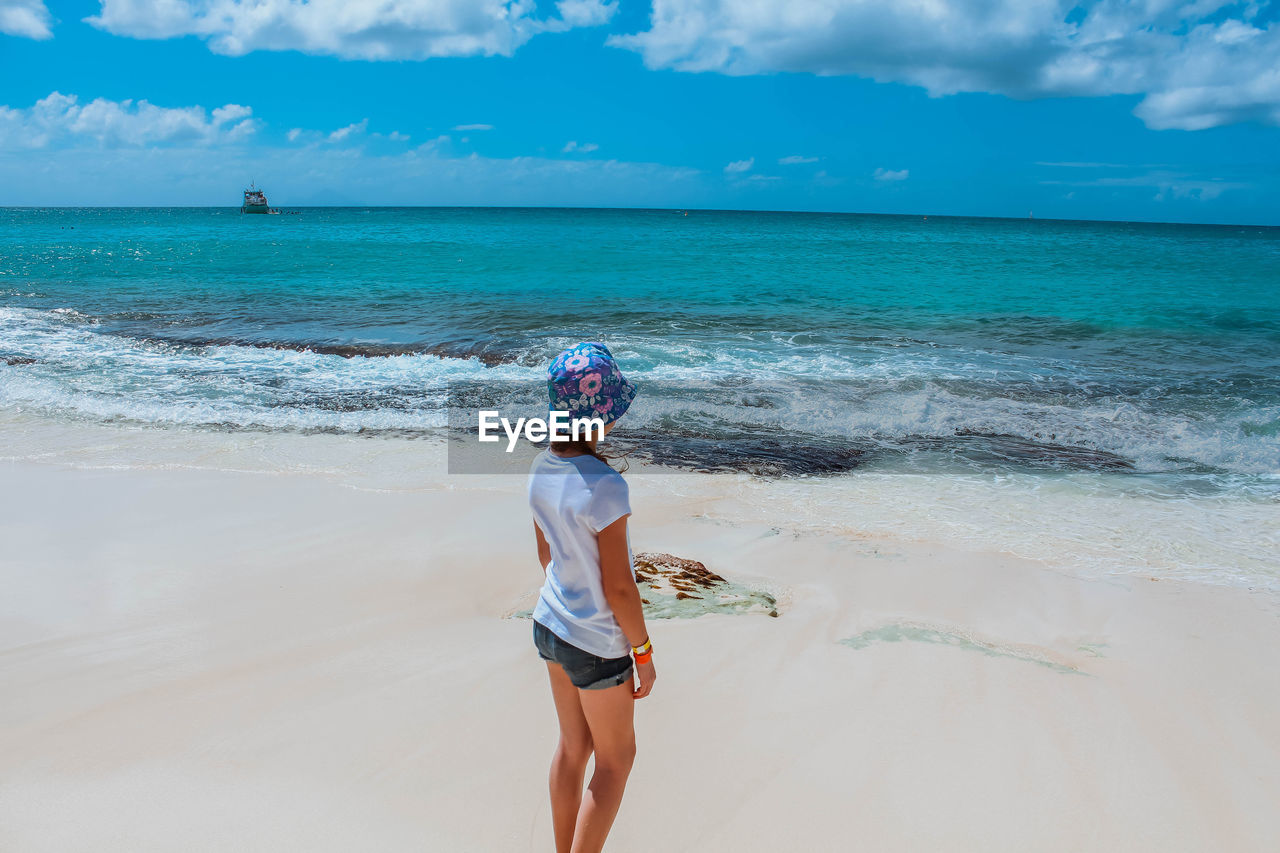 Rear view of girl wearing hat while standing at beach against cloudy sky