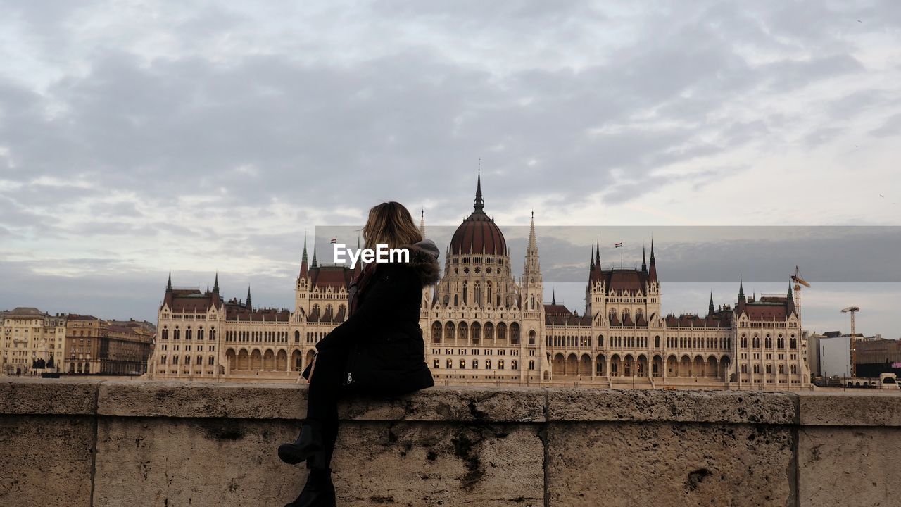 Woman sitting on retaining wall against hungarian parliament building