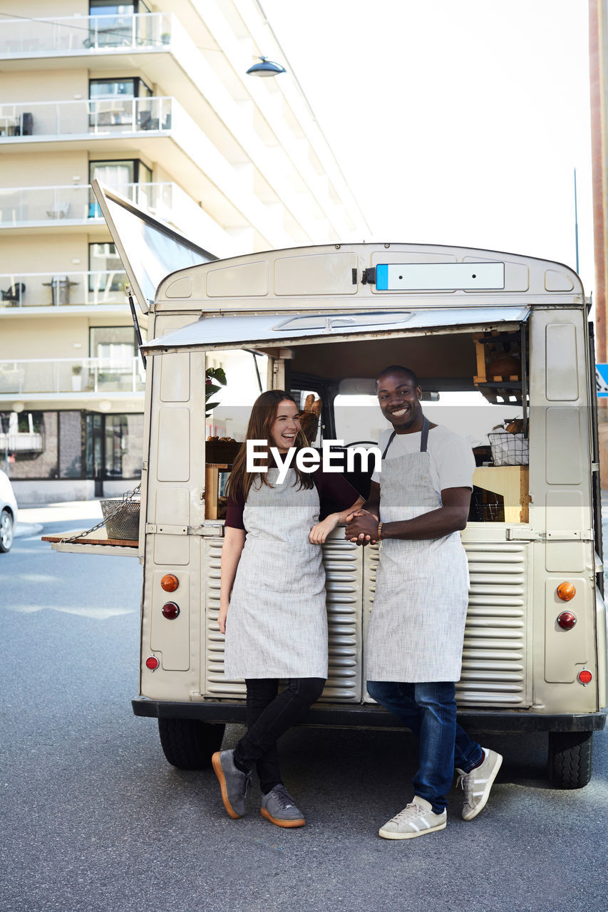 Full length of smiling male and female owners standing outside food truck parked on city street