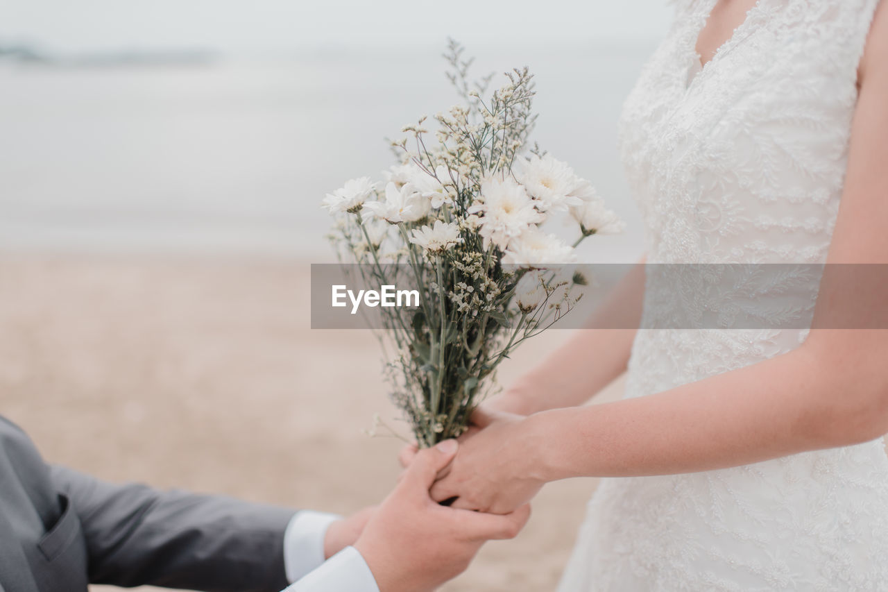 Midsection of groom holding hands of bride with bouquet at beach