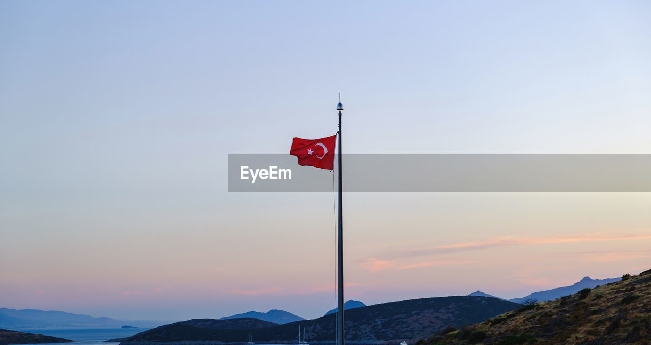 flag, sky, patriotism, mountain, environment, nature, red, scenics - nature, beauty in nature, no people, mountain range, tranquility, water, sunset, outdoors, tranquil scene, landscape, wind, land, sea, pole, cloud, travel destinations, symbol, day, travel