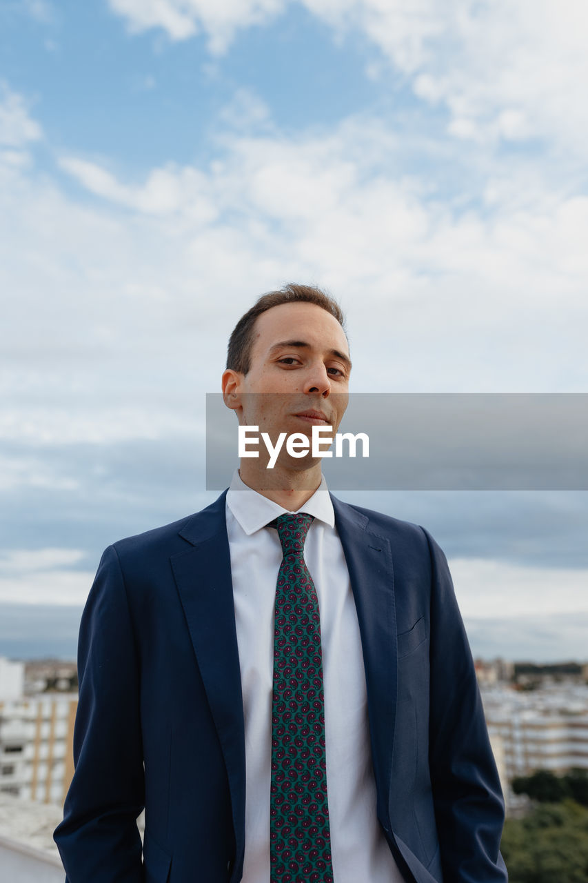 Young businessman in a suit and tie looking at camera and smiling while standing with a cloudy sky