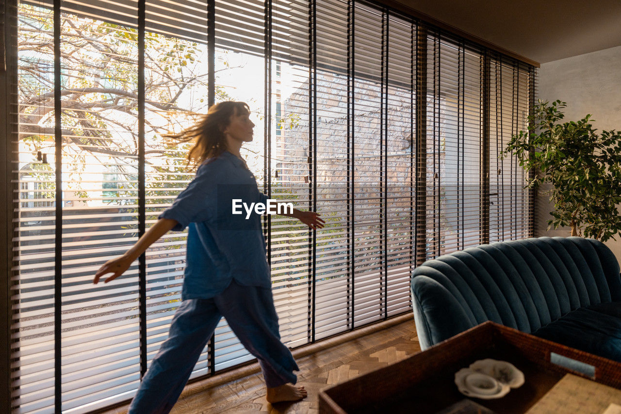 one person, adult, indoors, window covering, interior design, full length, women, window, lifestyles, standing, architecture, nature, day, home, casual clothing, side view, looking, furniture, young adult, room, hairstyle, house, sunlight, home interior, female, domestic life