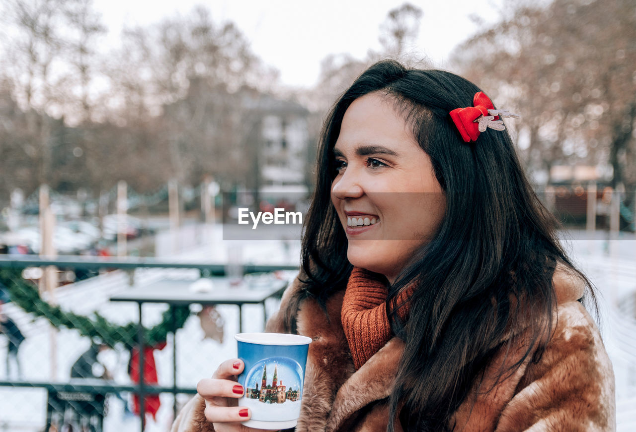 Portrait of happy beautiful young woman holding cup of mulled wine at christmas market in city