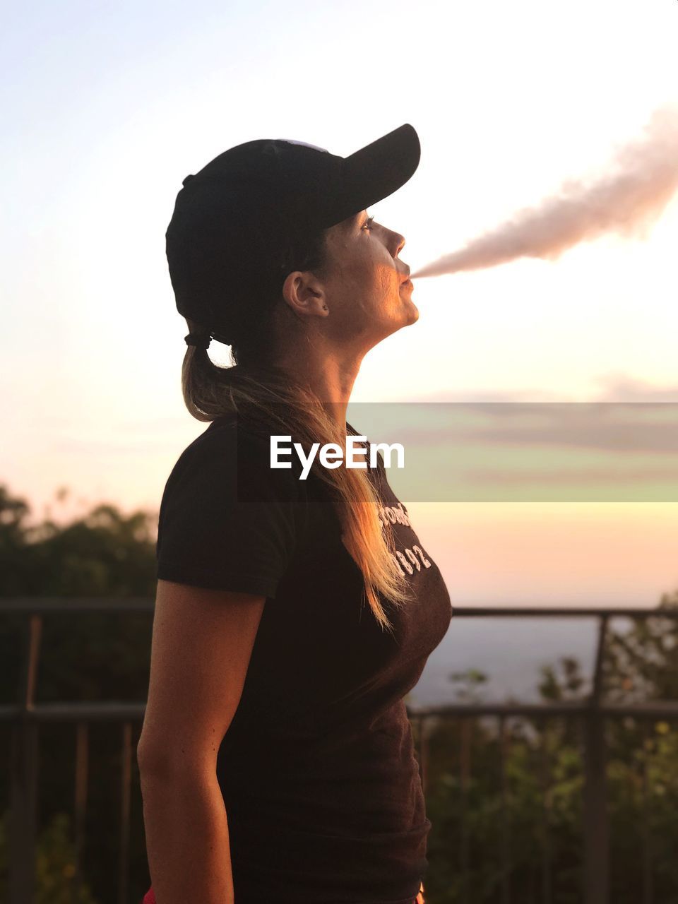 Side view of woman smoking while looking away against sky during sunset