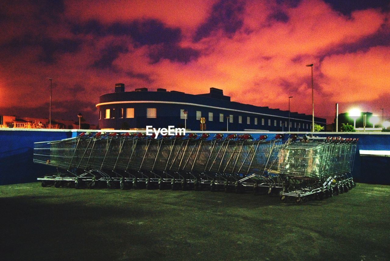 Shopping carts on field by building against sky during sunset