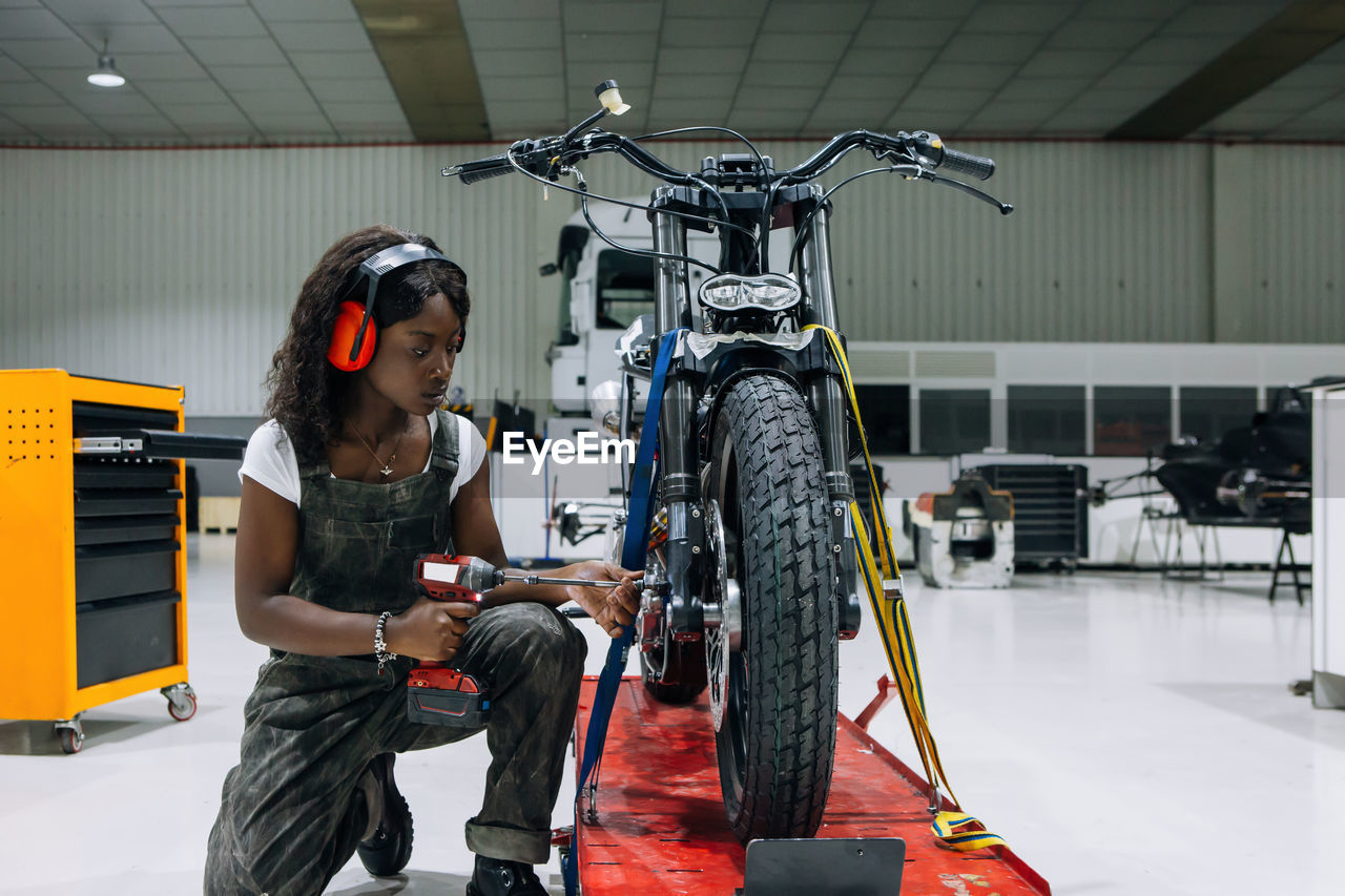Side view of skilled african american female mechanic in protective headphones using power screwdriver while assembling custom motorcycle in workshop