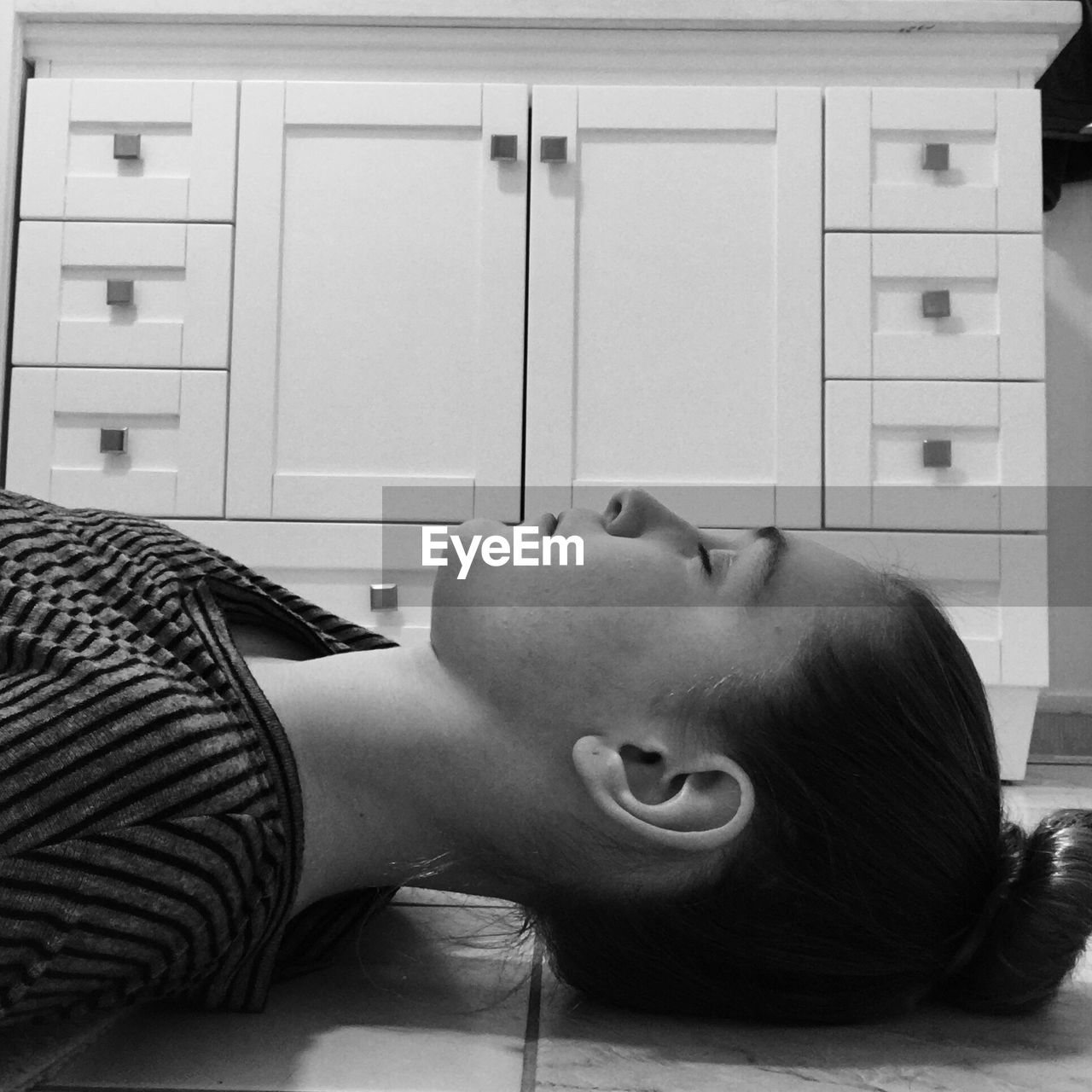 Side view of young woman lying on floor with eyes closed by cabinet at home