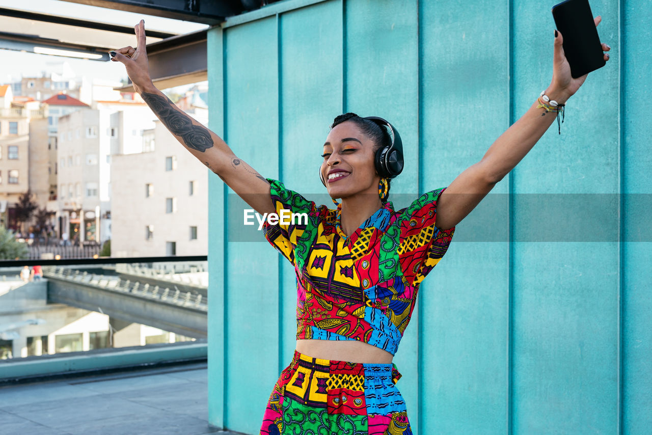 Cheerful ethnic woman in colorful clothes raising arms with smartphone and gesturing v sign while standing on terrace of modern building and listening to music in daytime