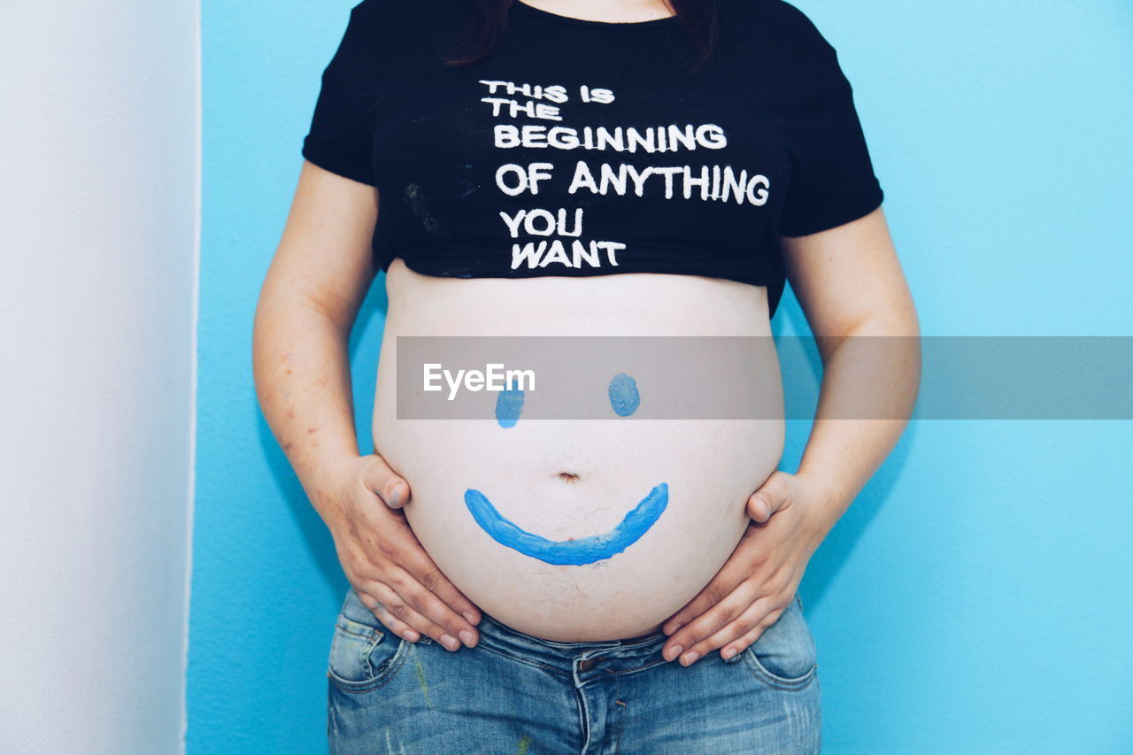 Midsection of pregnant woman with painted smile on his belly  against blue background