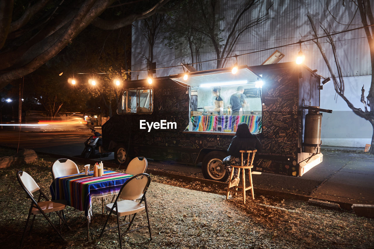 Table with chairs placed near client and food truck with workers illuminated by lamps at nighttime