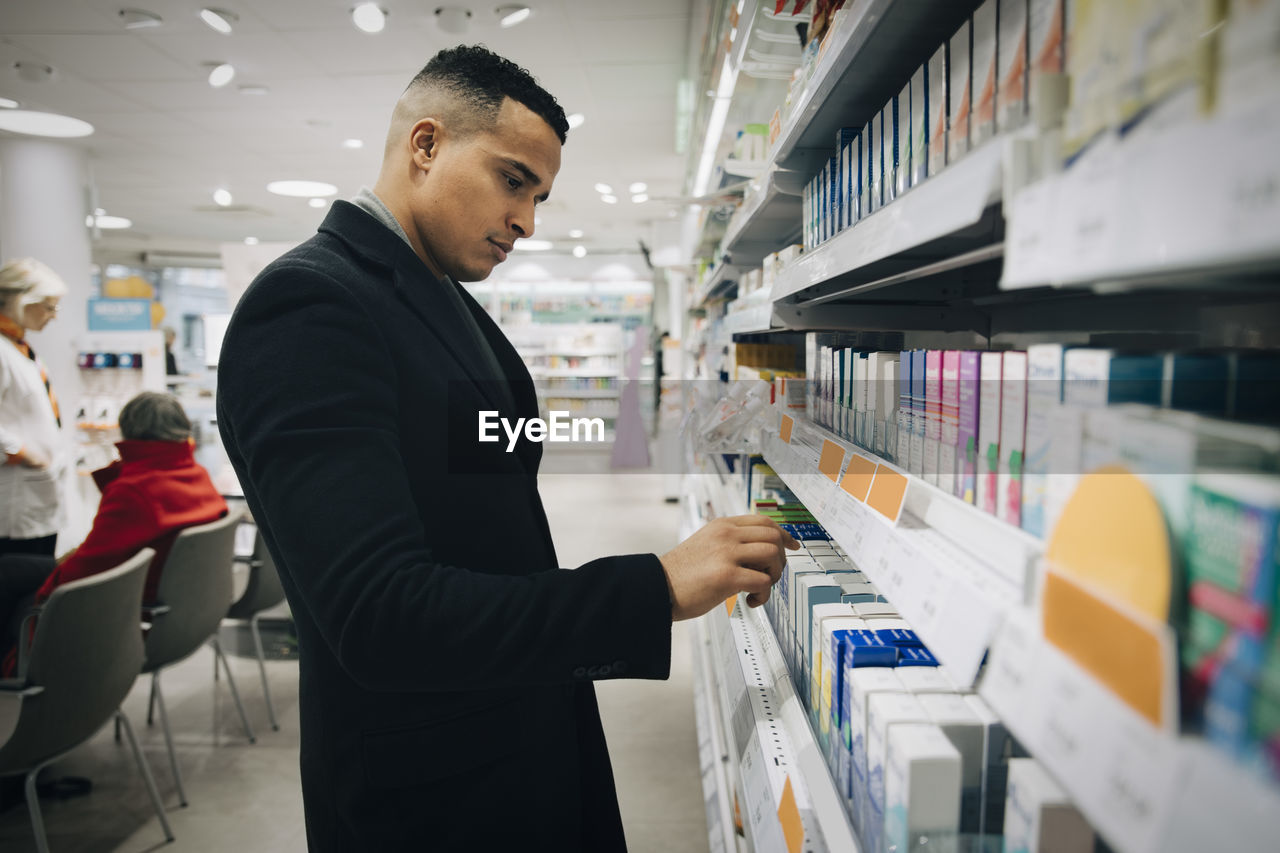 Side view of male customer choosing medicine from rack at pharmacy store