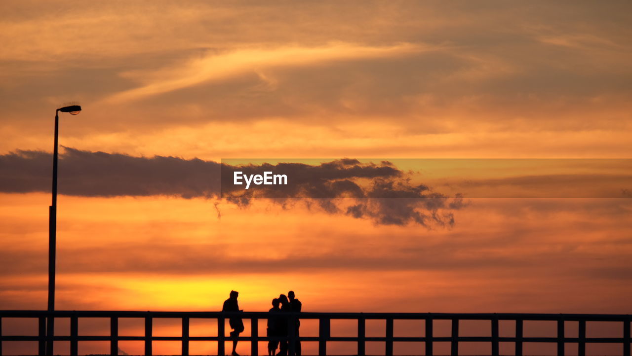 SILHOUETTE PEOPLE BY RAILING AGAINST SEA DURING SUNSET