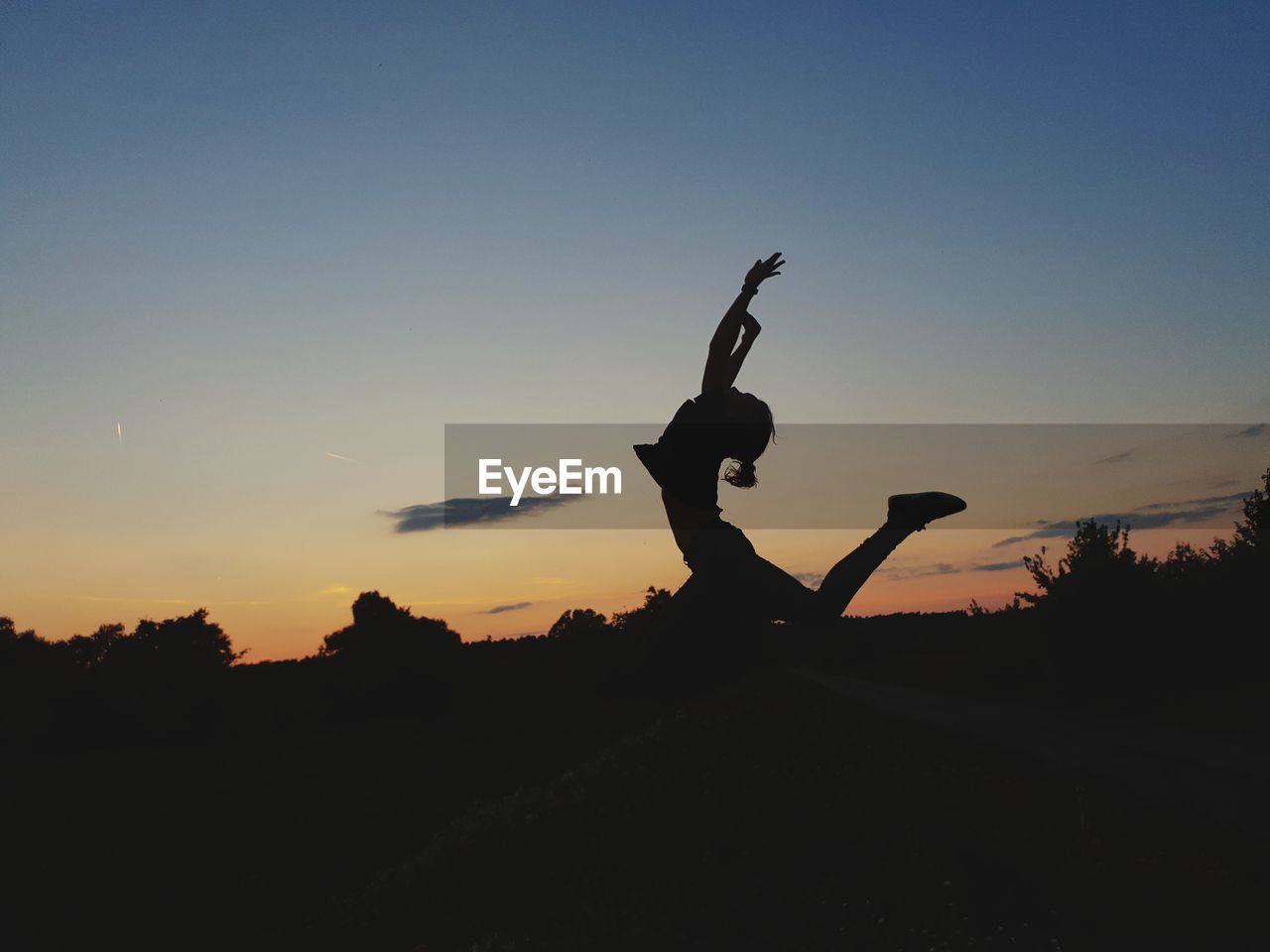 Silhouette young woman with arms raised jumping on field against sky during sunset