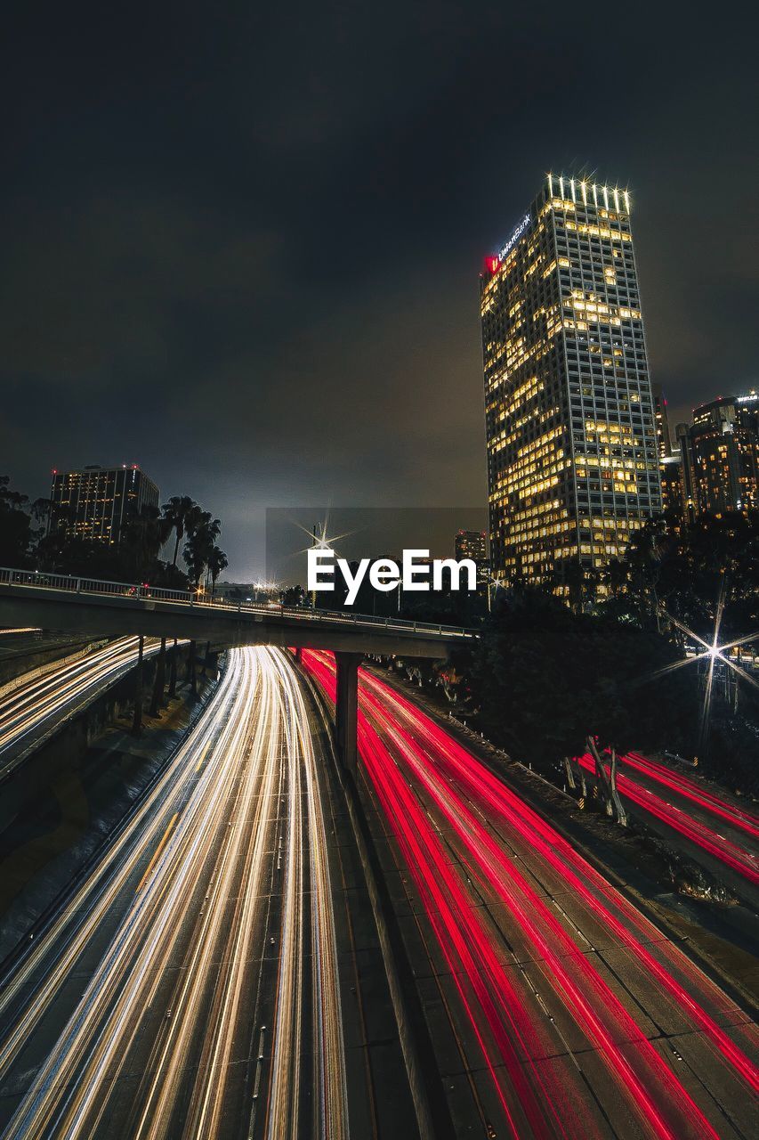 Light trails on road by illuminated buildings against sky at night