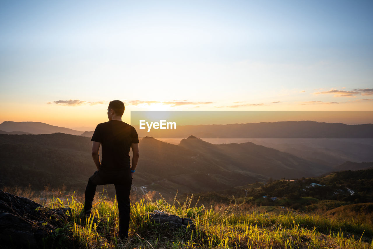 REAR VIEW OF MAN LOOKING AT MOUNTAINS AGAINST SUNSET SKY