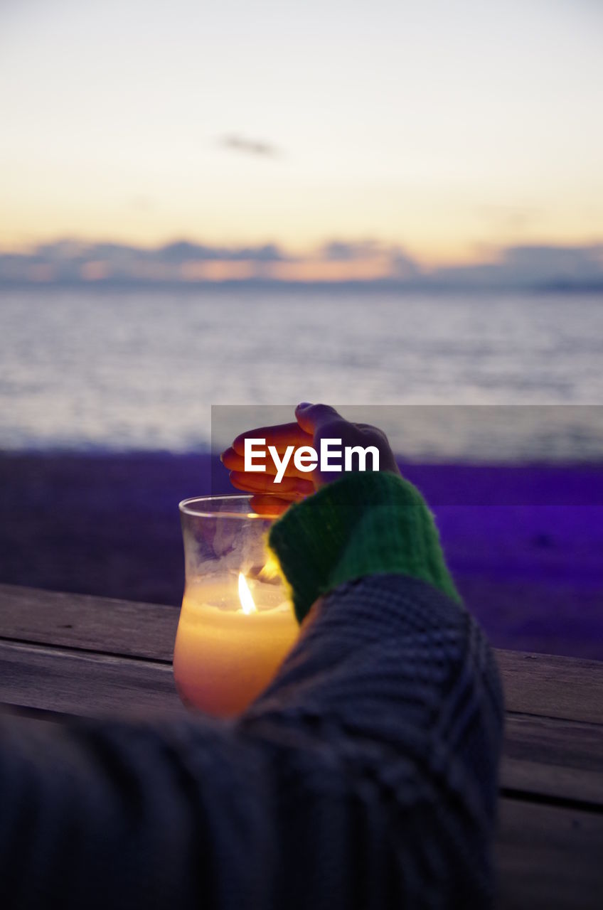 Cropped hand of person covering illuminated candle on table at beach