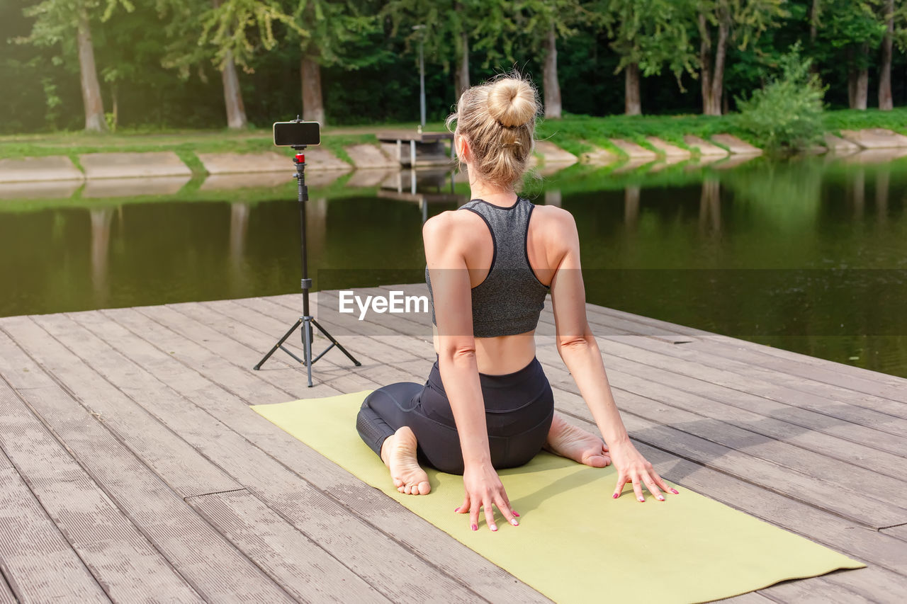 A woman on a wooden platform in summer, does yoga on green mat by pond
