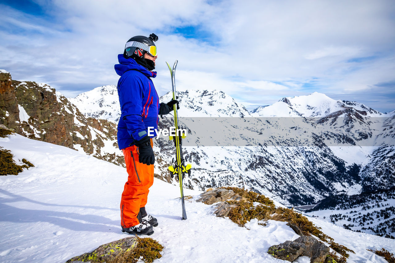 Man in ski-wear standing on snowcapped mountain against sky