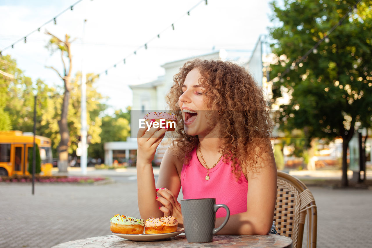 Young attractive curly woman sits at table in cafe on summer terrace. colorful delicious donuts,