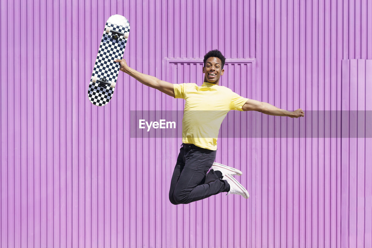 Cheerful man with arms outstretched holding skateboard whole jumping in purple corrugated iron