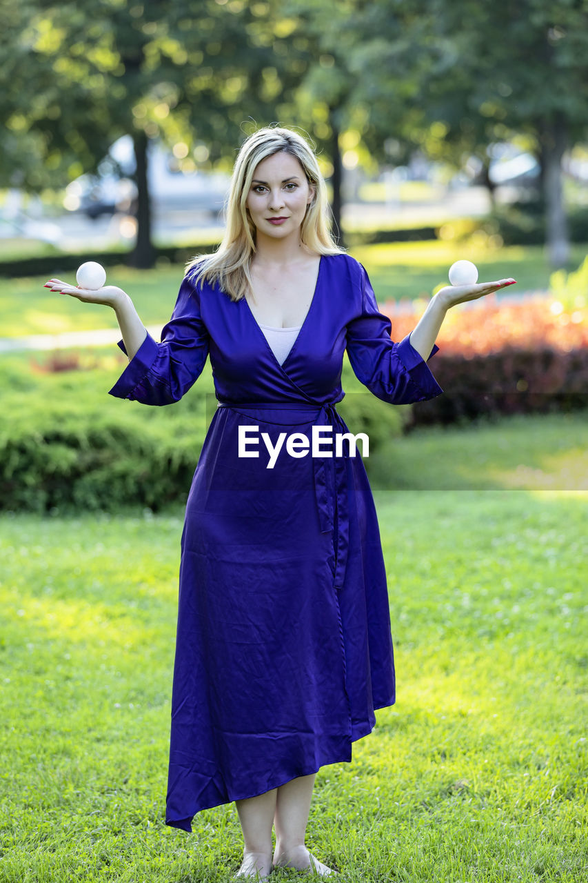 Beautiful blonde in purple dress is balancing in public park and holding a small balls