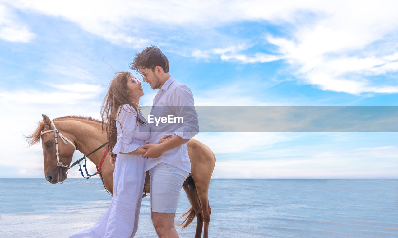 Side view of couple standing by horse on beach against sky