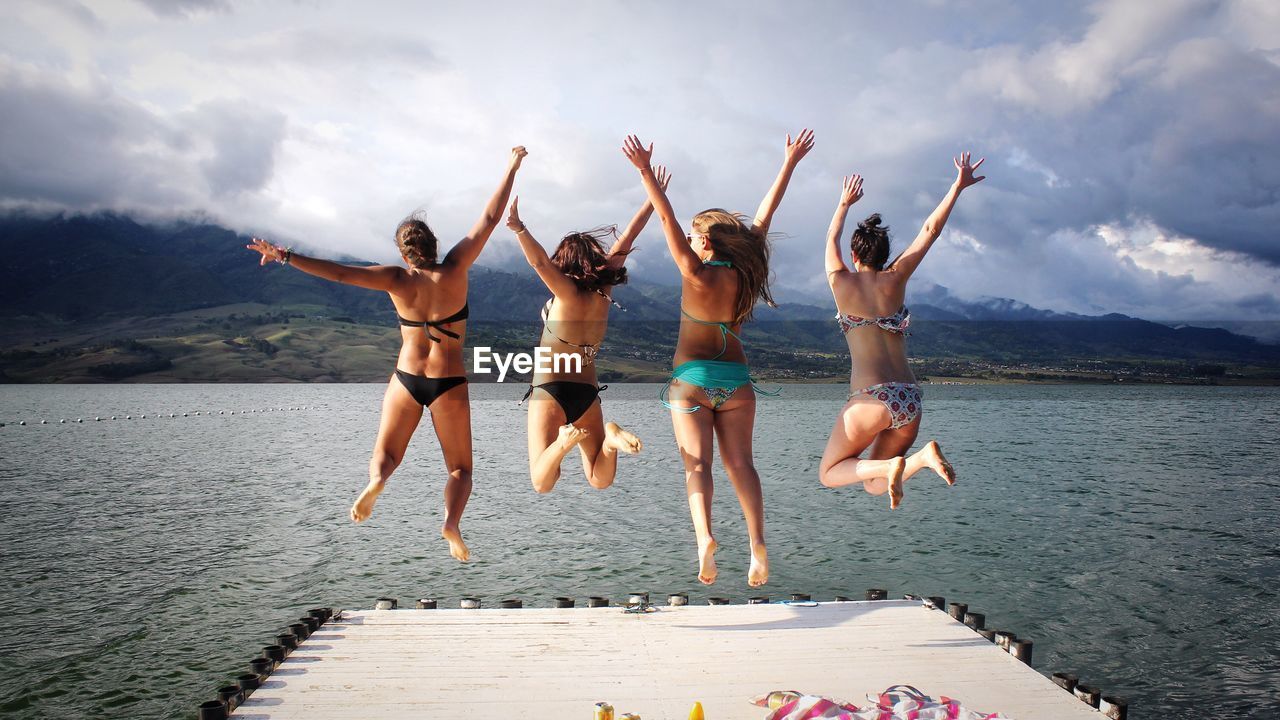 Rear view of women in bikini jumping in lake against sky