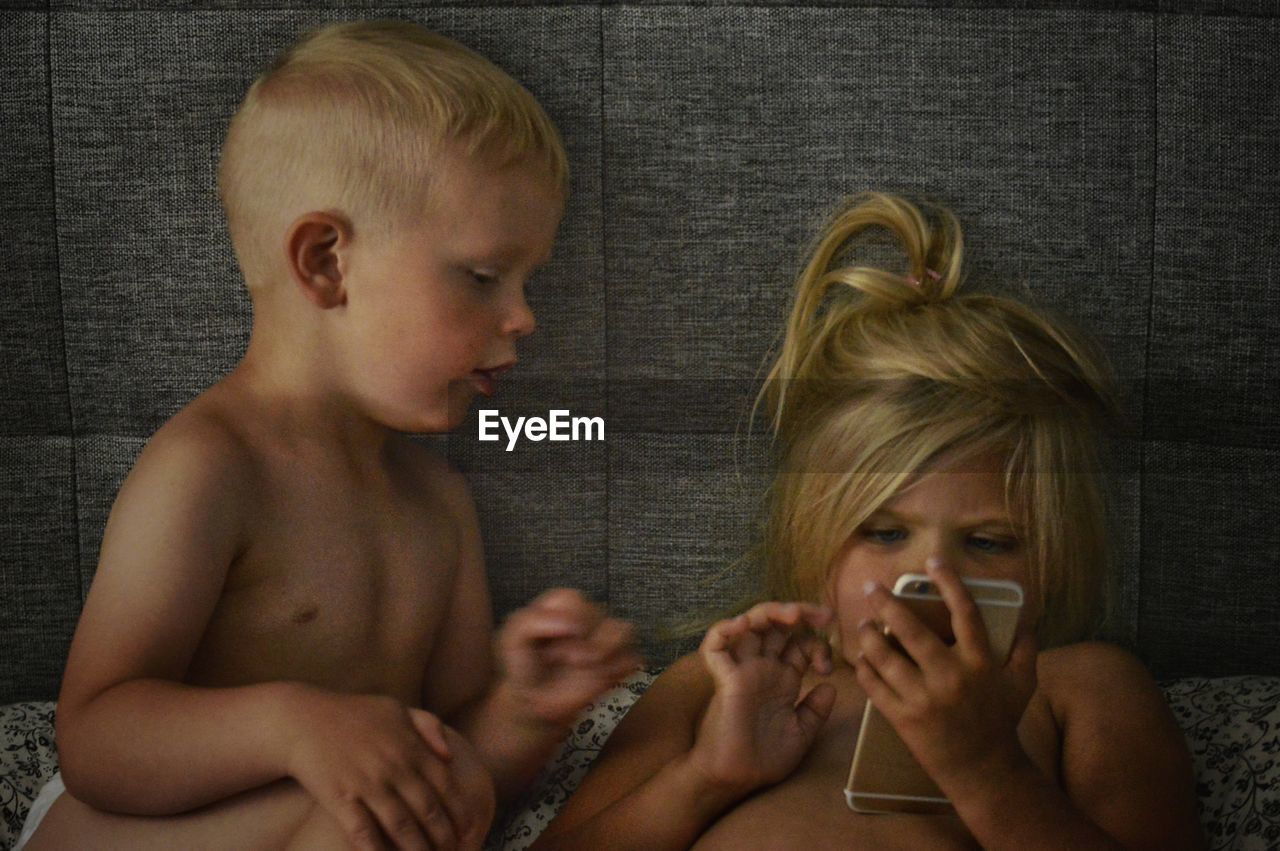 Shirtless boy looking at sister using smart phone on bed