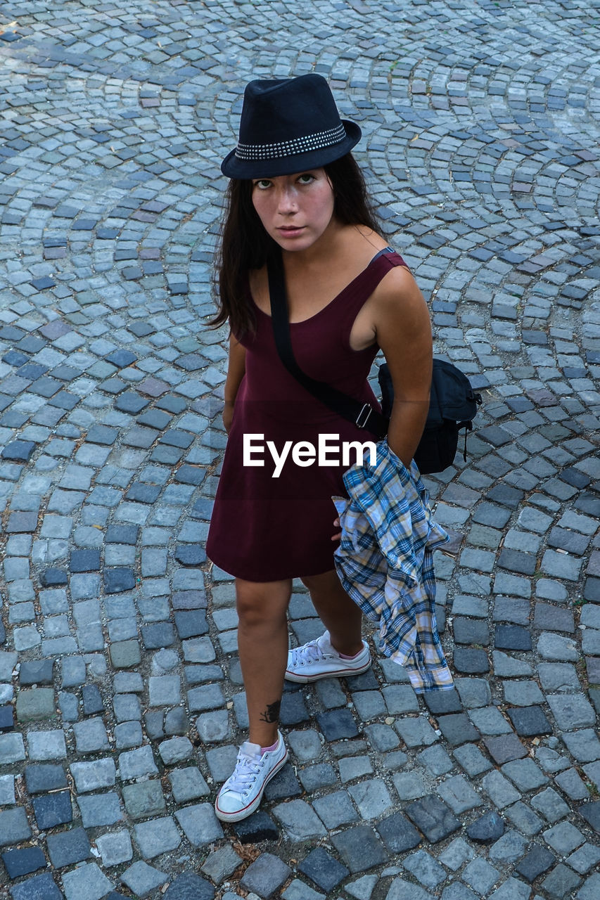 A young brunette woman in a burgundy dress and a black hat stands on the pavement road