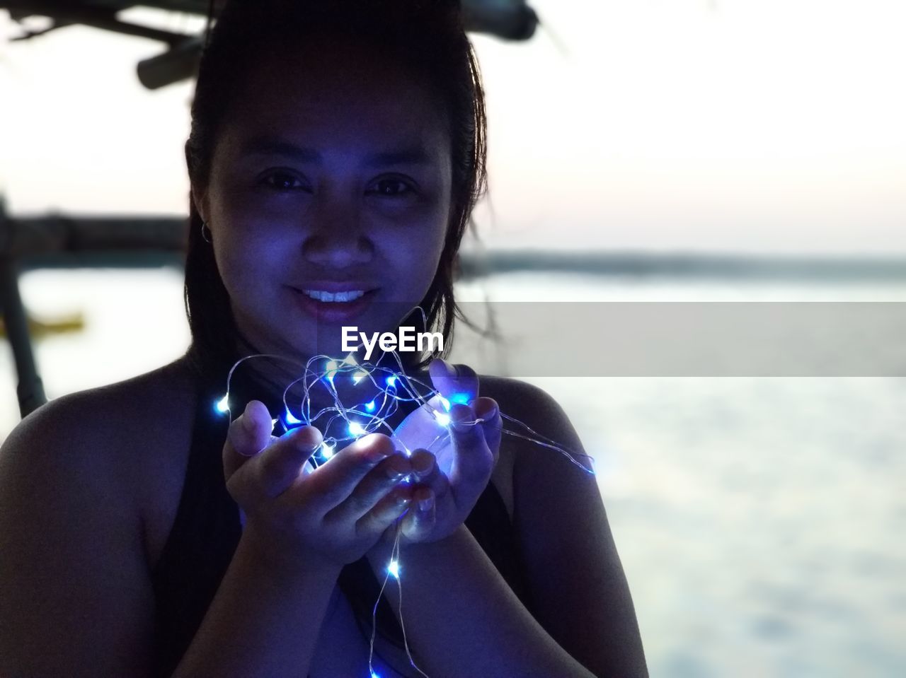 Close-up portrait of young woman with illuminated string lights against lake