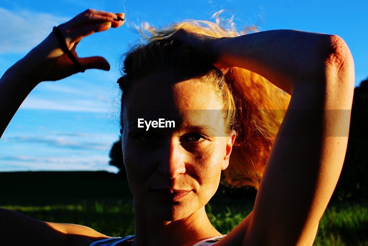 Portrait of young woman tying hair against sky during sunset