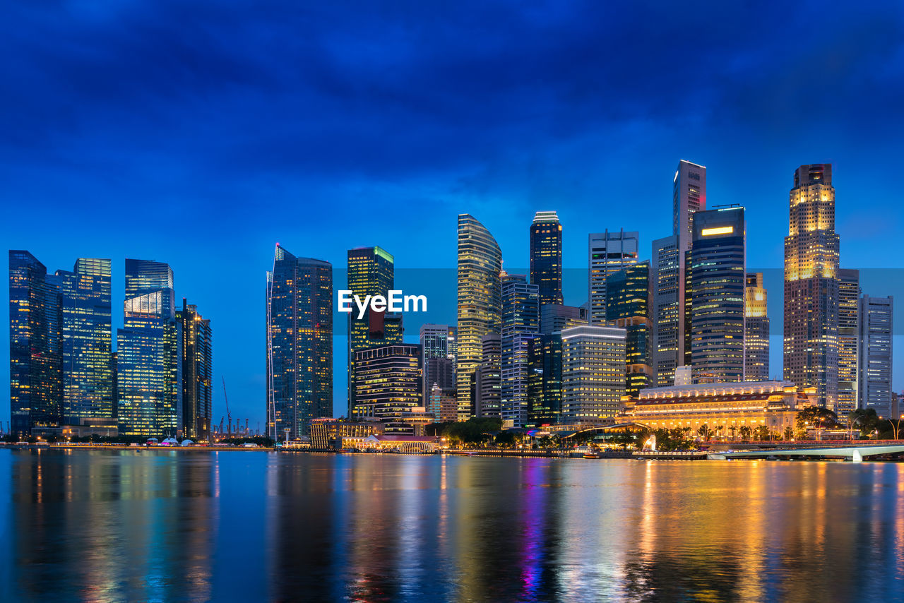 Illuminated modern buildings by bay against blue sky