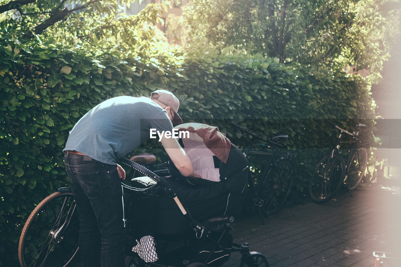 Full length of father bending over baby carriage on footpath at park during sunny day