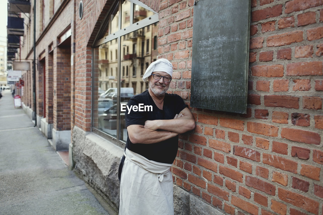 Portrait of senior male baker leaning on brick wall outside bakery