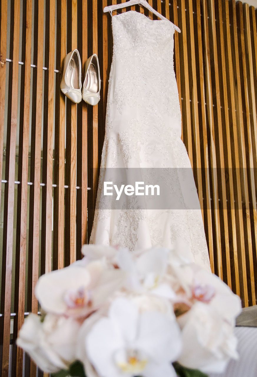 Wedding dress hanging on wood next to shoes with bouquet flowers out of focus in the foreground