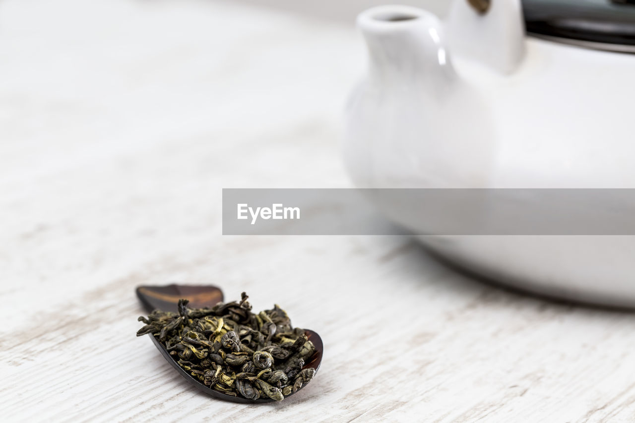 Green tea in a wooden spoon is on a table in front of a kettle and a cup in a tea room.