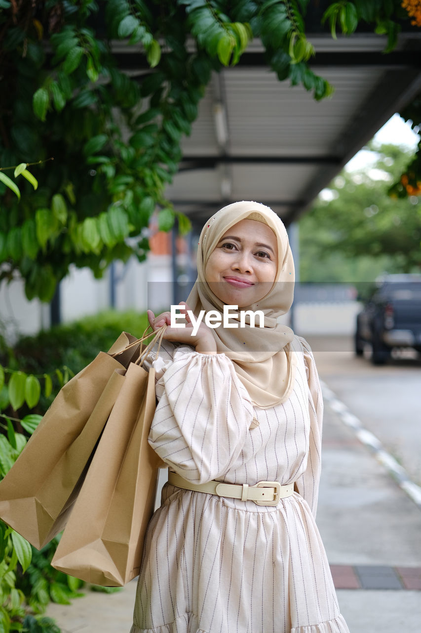 Young woman standing against shopping mall after shopping while holding paper bag 