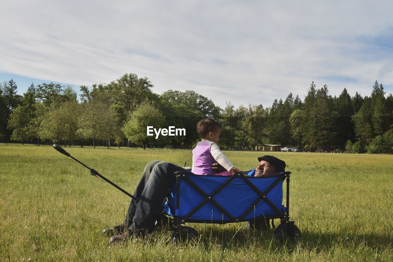 Father and daughter in cart on grass against trees and sky