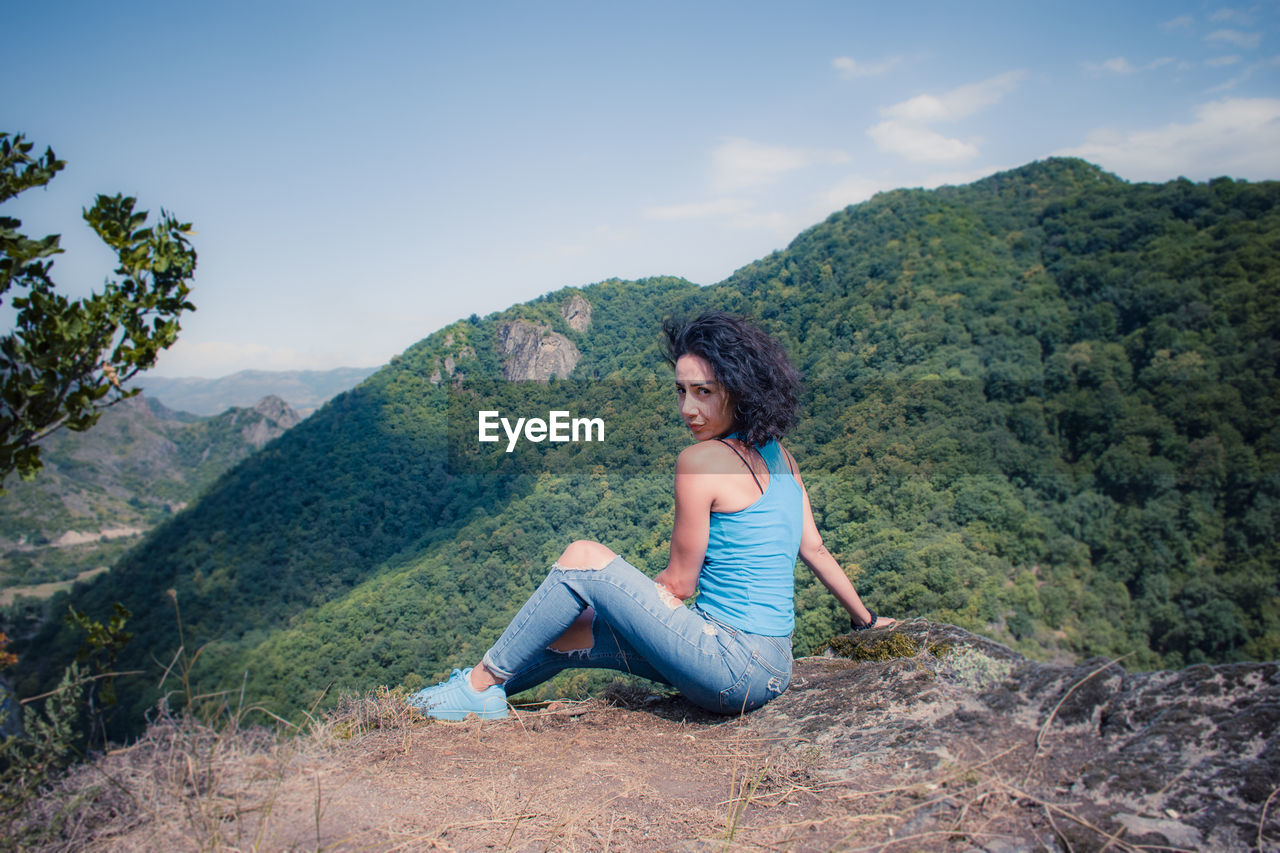 BEAUTIFUL YOUNG WOMAN SITTING ON MOUNTAIN AGAINST SKY